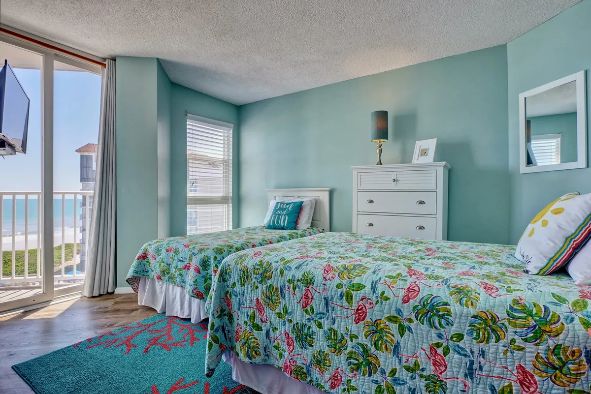 A cheerful guest bedroom with two twin beds covered in colorful quilts, a large window revealing a beach view, a decorative lamp on the nightstand, and a dresser with a mirror. The space feels bright, airy, and family-friendly.