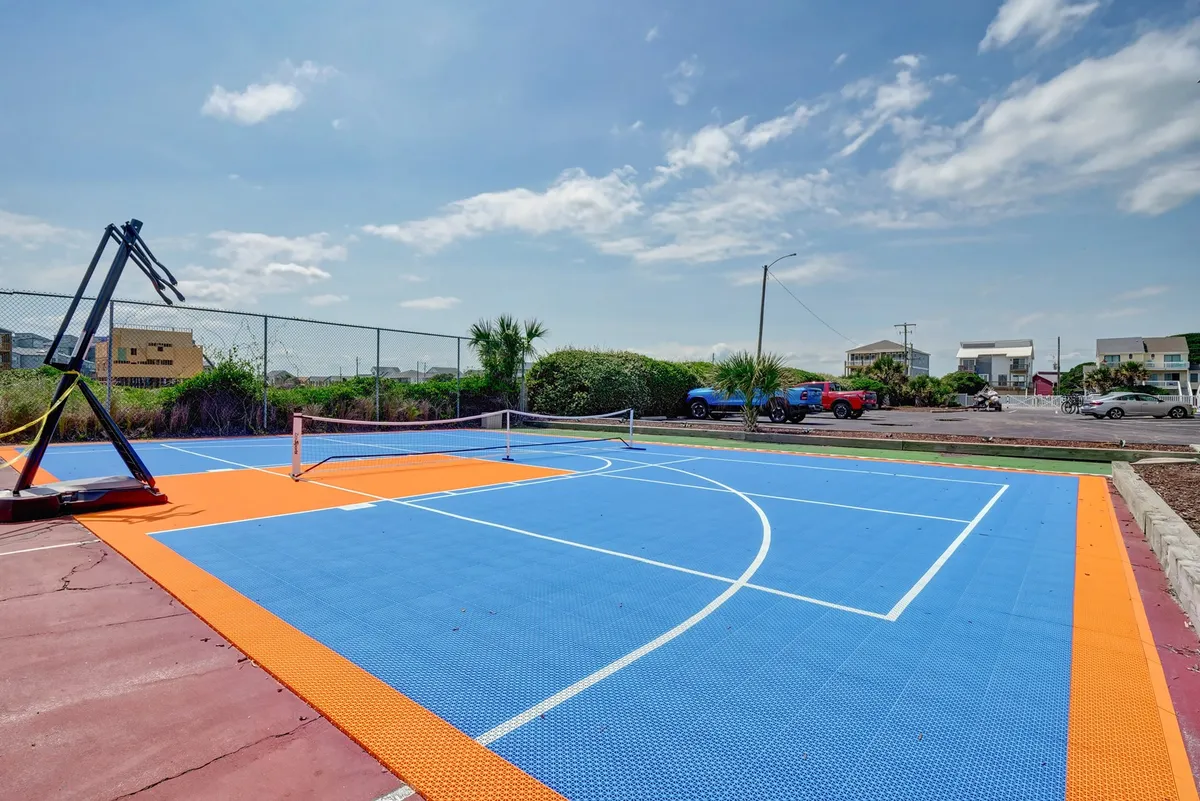 A vibrant outdoor sports area featuring a basketball court and a tennis court surrounded by lush plants and nearby houses. The courts are clean and well-maintained, offering a sense of energetic outdoor recreation and community.