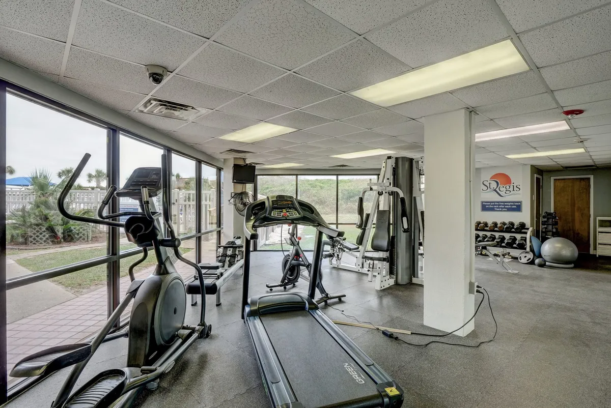 A fully equipped fitness center featuring treadmills, elliptical machines, weight equipment, and natural light filtering through large windows. The space feels motivational and active, suitable for group fitness or personal training.