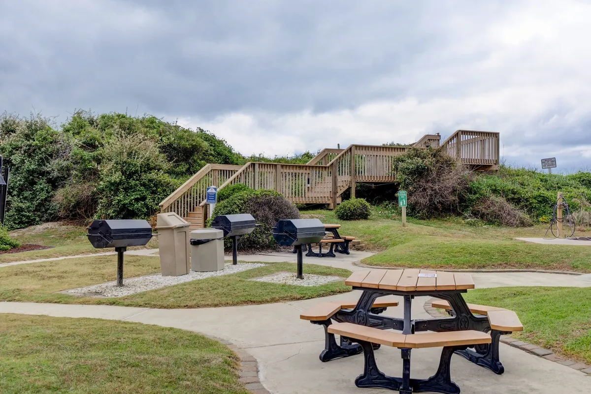 An inviting outdoor grilling area set among picnic tables, with winding paths and greenery creating a welcoming communal dining space. The scene suggests relaxed gatherings and festive cookouts.