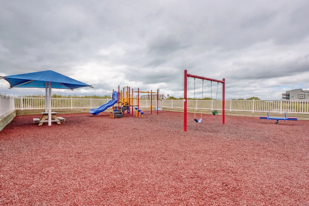 A colorful playground equipped with swings, slides, and a picnic table shaded by a sunshade. Families are seen enjoying picnics while children play, capturing youthful energy and community spirit.
