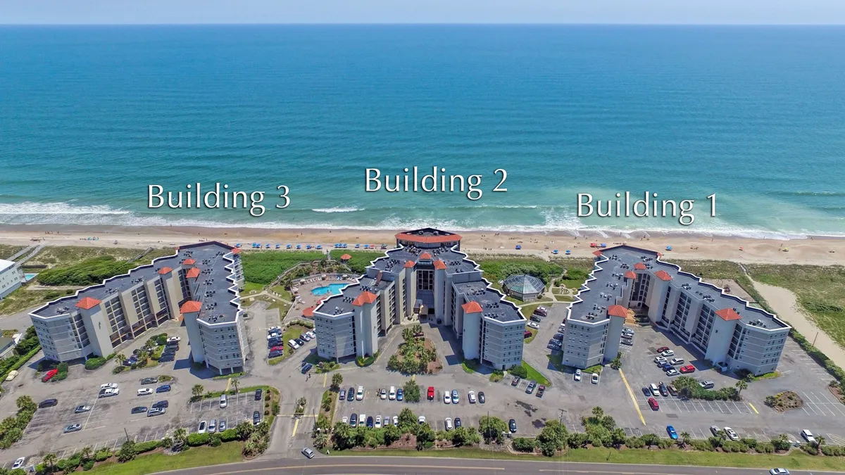 This exterior image features three modern buildings surrounding a spacious swimming pool with a striking ocean view in the background. The scene captures guests swimming, sunbathing by the pool, and the inviting expanse of the beach just beyond the resort. The setting is tranquil and open, reflecting the seamless connection between the resort's amenities and its spectacular coastal location.
