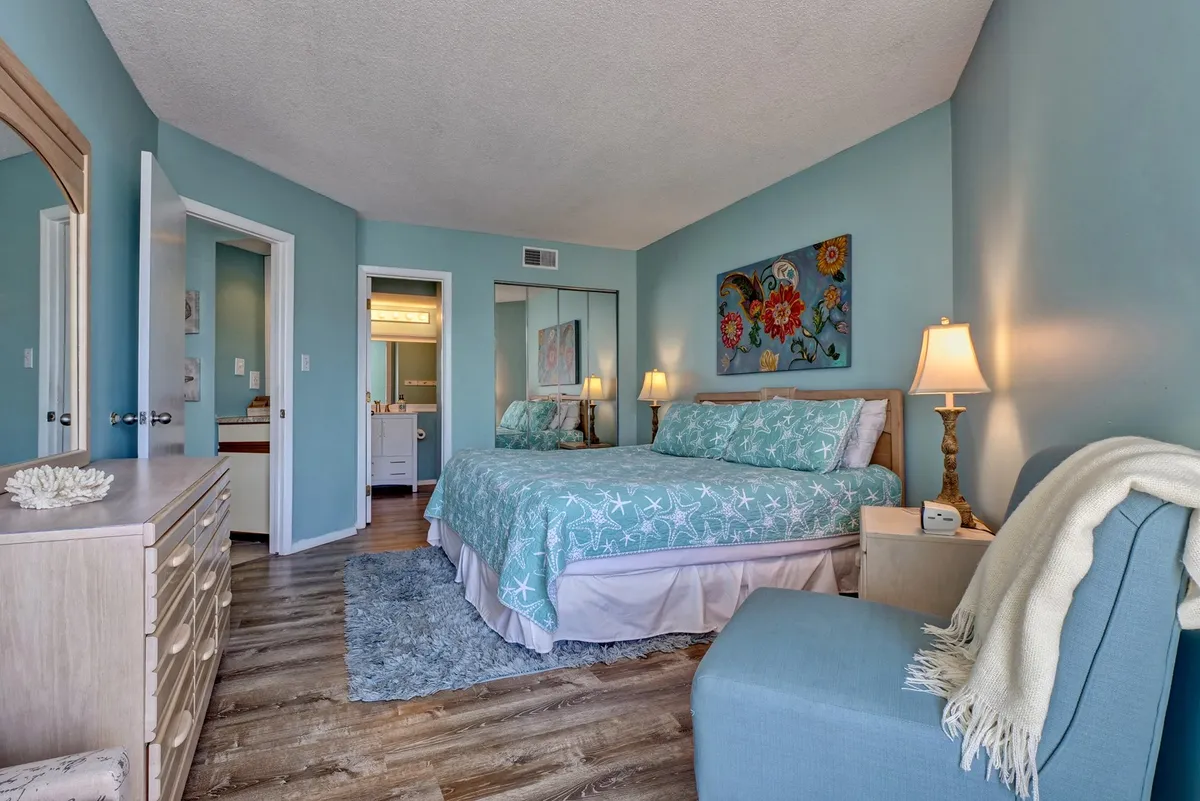 This image features a cozy bedroom highlighted by turquoise bedding, decorative artwork on the wall, and a nightstand with a lamp. The space is awash with natural light, emphasizing a tranquil and refreshing atmosphere ideal for relaxation.