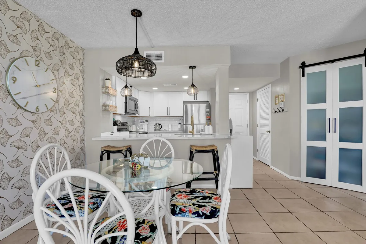A glass dining table sits beneath a contemporary pendant light, set against a decorative wall with a cheerful leaf pattern. The dining area is adjacent to a modern kitchen, creating an inviting space for meals and casual conversations among family and friends.