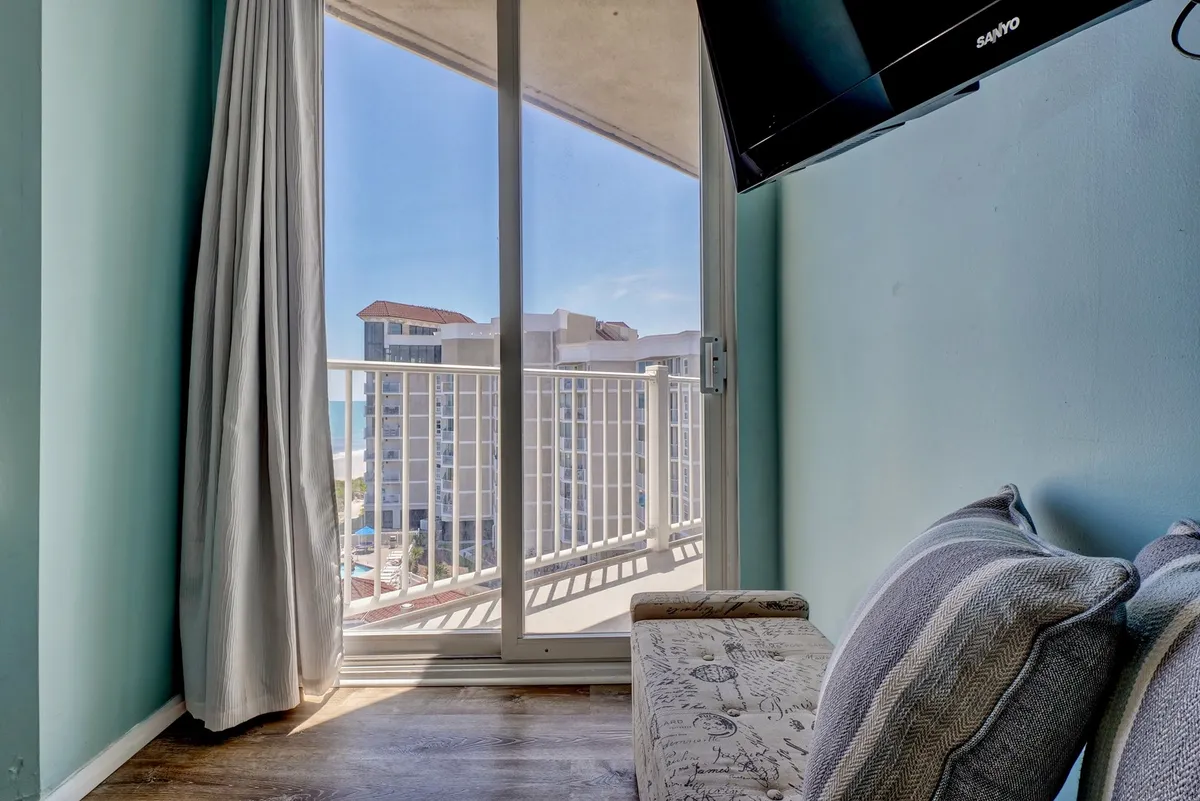 A tranquil living area with a sofa positioned to enjoy direct views through glass doors onto a private balcony. The ocean is visible in the background, offering a seamless transition between indoor comfort and outdoor serenity. Soft furnishings and light curtains enhance the relaxed, cozy vibe.