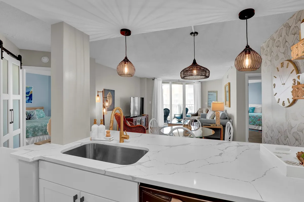 This image features an open-concept space that seamlessly connects a modern kitchen island, dining area, and living room seating. Natural light brightens the area, and coastal decorative touches are visible, such as light-colored furniture and a decorative wall clock. The overall layout is airy and welcoming, perfect for gatherings or relaxation.
