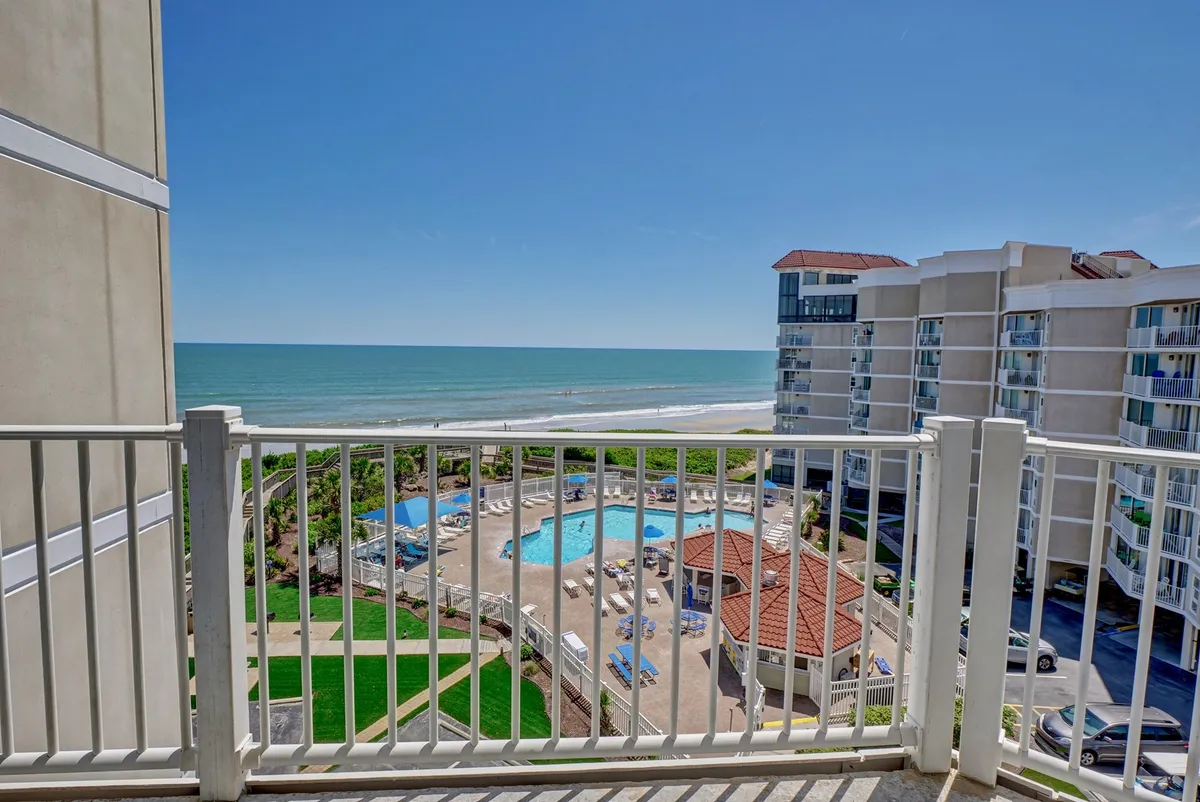 A sweeping balcony perspective with a panoramic view of the ocean, swimming pool, lush greenery, and nearby buildings. The image captures the tranquil blue of the Atlantic and the inviting resort amenities below.