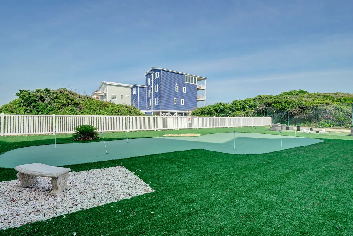 This image highlights a well-manicured putting green bordered by lush landscaping, with a stone bench and a blue house nearby. The area is serene and inviting, hinting at leisurely outdoor activities in a resort setting.