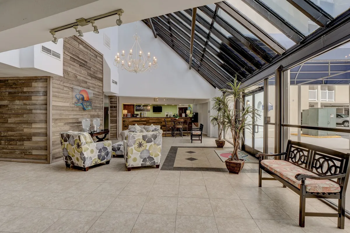 A welcoming interior lounge area featuring a chandelier, a reception desk, comfortable seating, large glass windows, and lush indoor plants. Sunlight streams through the windows, creating a warm and relaxed atmosphere ideal for socializing, check-ins, or casual meetings.