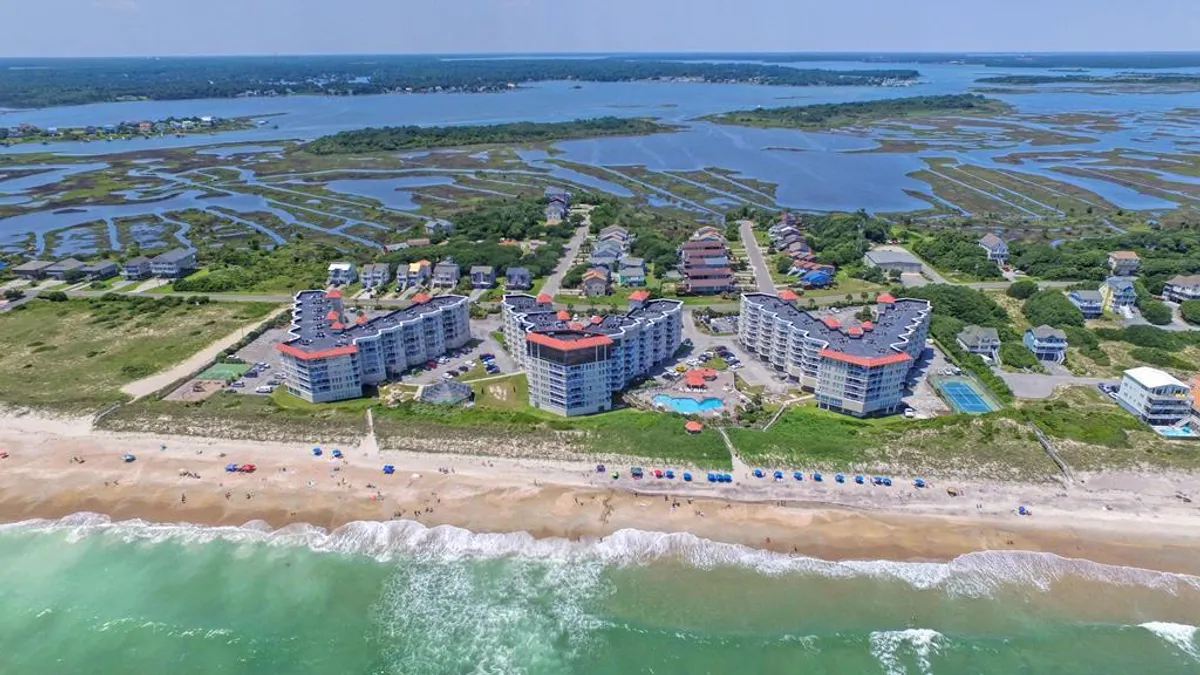 This exterior image captures the beachfront buildings of the resort complex, a large swimming pool, a sandy beach, gentle ocean waves, and stretches of marshland in the background. The pool is surrounded by lounge chairs, emphasizing opportunities for swimming and sunbathing, while the beachfront location hints at direct access to scenic walking and jogging paths.