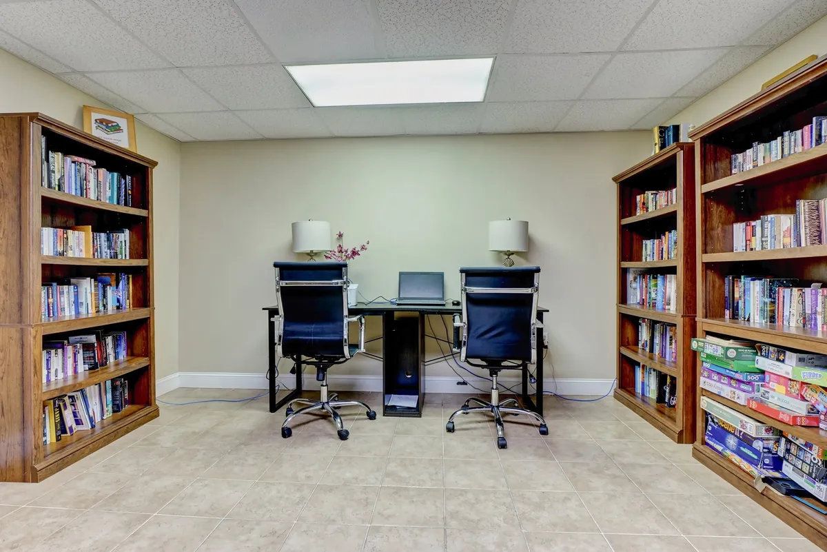 A bright interior space filled with bookshelves, desks, and computers, offering both private and collaborative workstations. Warm lighting and comfortable chairs create an inviting environment for focused work, reading, or meetings. The design is both practical and welcoming, suggesting a productive atmosphere for remote workers or business retreats.