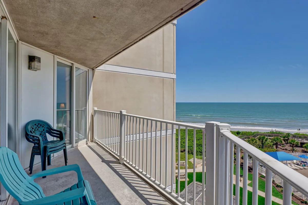This exterior shot captures the oceanfront balcony with blue chairs and sturdy railings, offering expansive views of the water. The space is inviting for morning coffee, evening cocktails, or peaceful reflection while taking in the rhythmic waves.